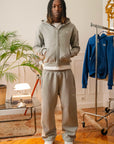 Homme debout portant un ensemble streetwear Cynique en coton 450 GSM : hoodie zippé gris clair avec logo brodé blanc et jogging assorti coupe oversize. Intérieur au style rétro.