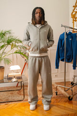 Homme debout portant un ensemble streetwear Cynique en coton 450 GSM : hoodie zippé gris clair avec logo brodé blanc et jogging assorti coupe oversize. Intérieur au style rétro.
