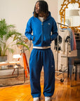 Hoodie zippé bleu électrique et jogging assorti Cynique portés par un mannequin dans un intérieur vintage. Ensemble unisexe en coton 450 GSM, coupe oversize, logo brodé blanc sur le cœur et la cuisse.