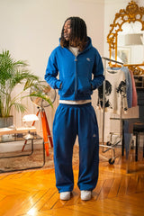 Hoodie zippé bleu électrique et jogging assorti Cynique portés par un mannequin dans un intérieur vintage. Ensemble unisexe en coton 450 GSM, coupe oversize, logo brodé blanc sur le cœur et la cuisse.