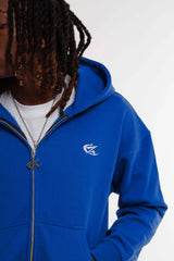 Gros plan sur hoodie zippé bleu Cynique porté ouvert, avec logo brodé blanc côté cœur, zip métal et t-shirt blanc en dessous. Coton épais 450 GSM, coupe oversize.