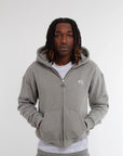 Homme portant un hoodie zippé gris clair Cynique, coton 450 GSM, coupe oversize unisexe. Logo brodé blanc sur la poitrine, poches kangourou, capuche doublée et zip métal. Style streetwear épuré.