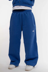 Jogging bleu électrique Cynique en coton épais 450 GSM, coupe oversize avec cordons de serrage aux chevilles. Logo brodé blanc sur la cuisse droite. Porté avec sneakers blanches.