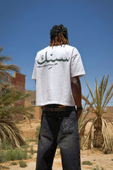 Vue arrière du t-shirt Cynique Maroc porté dans le désert. Lettrage arabe et slogan “damn all to crown.” Style affirmé et influence nord-africaine forte.