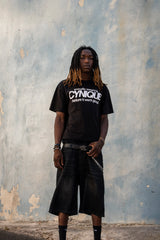 Look streetwear Cynique : T-shirt noir “proudly wearing Cynique before it went global.”, short en denim noir, ceinture logo métal. Photo urbaine sur fond de mur brut texturé.