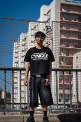 T-shirt streetwear Cynique noir “proudly wearing Cynique before it went global.” et jort denim noir. Look urbain assumé, photo en extérieur devant immeubles et grillage, ambiance raw.