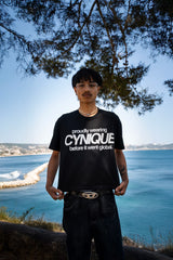 T-shirt noir Cynique “proudly wearing Cynique before it went global.” porté avec jean large, look streetwear minimaliste. Photo en extérieur, ambiance bord de mer.