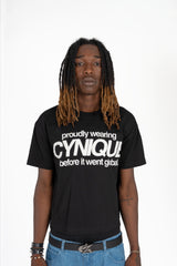 T-shirt noir Cynique “proudly wearing Cynique before it went global.” porté avec jean large bleu et ceinture logo métal. Look streetwear assumé, fond studio blanc.