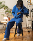 Homme assis sur un tabouret portant un ensemble hoodie zippé et jogging bleu électrique Cynique, coupe oversize, coton 450 GSM, logo brodé blanc, décor rétro intérieur.