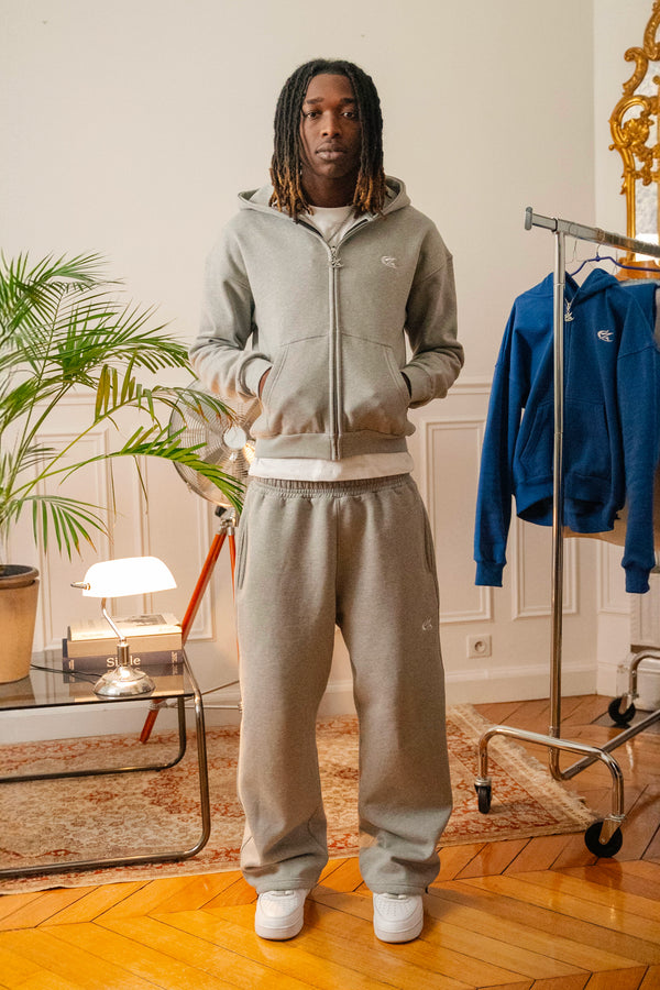 Male model wearing full Cynique Basic Gray tracksuit — zip hoodie and oversized joggers styled with white sneakers in modern indoor setting