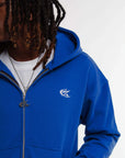 Gros plan sur hoodie zippé bleu Cynique porté ouvert, avec logo brodé blanc côté cœur, zip métal et t-shirt blanc en dessous. Coton épais 450 GSM, coupe oversize.