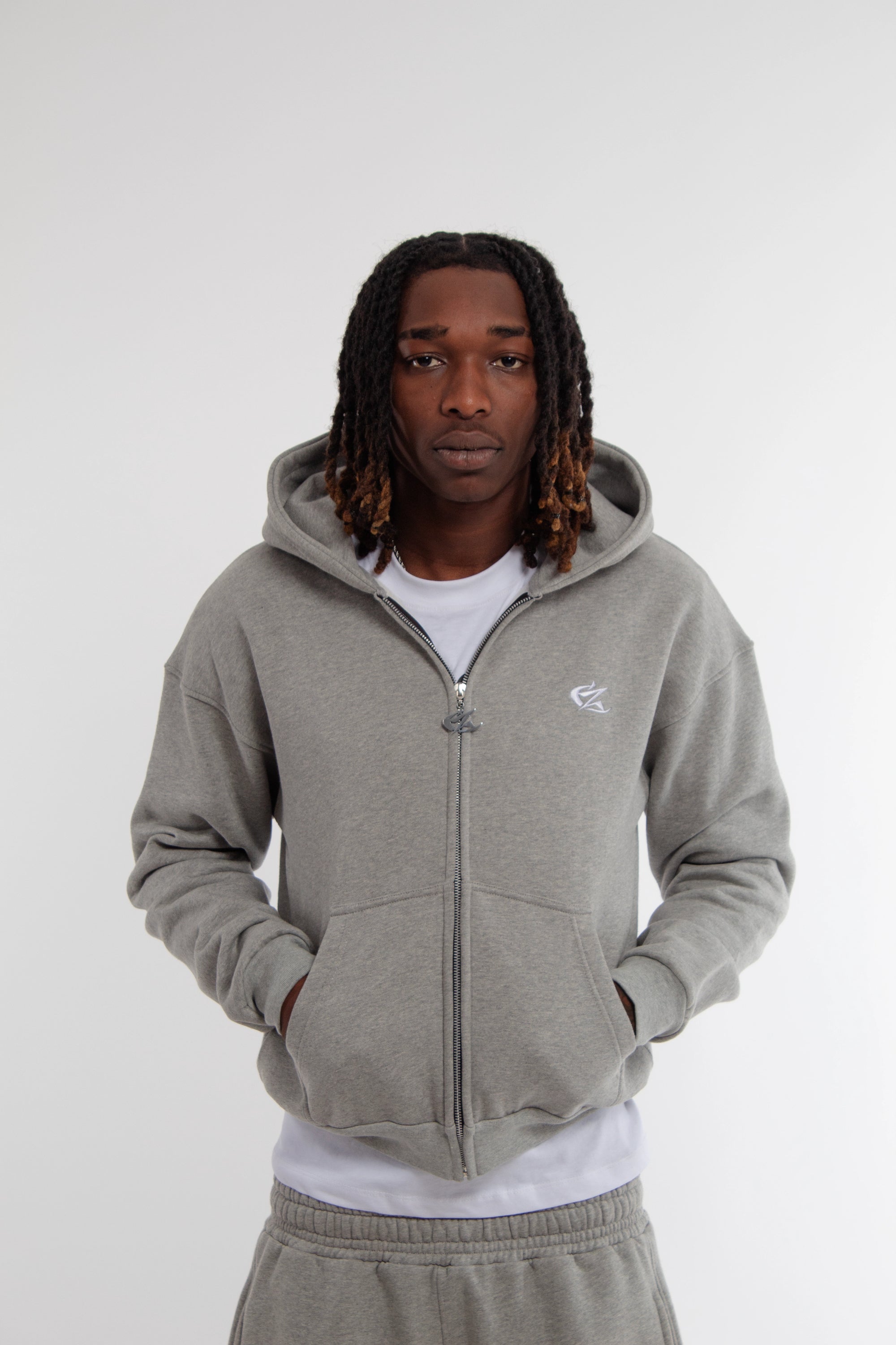 Homme portant un hoodie zippé gris clair Cynique, coton 450 GSM, coupe oversize unisexe. Logo brodé blanc sur la poitrine, poches kangourou, capuche doublée et zip métal. Style streetwear épuré.