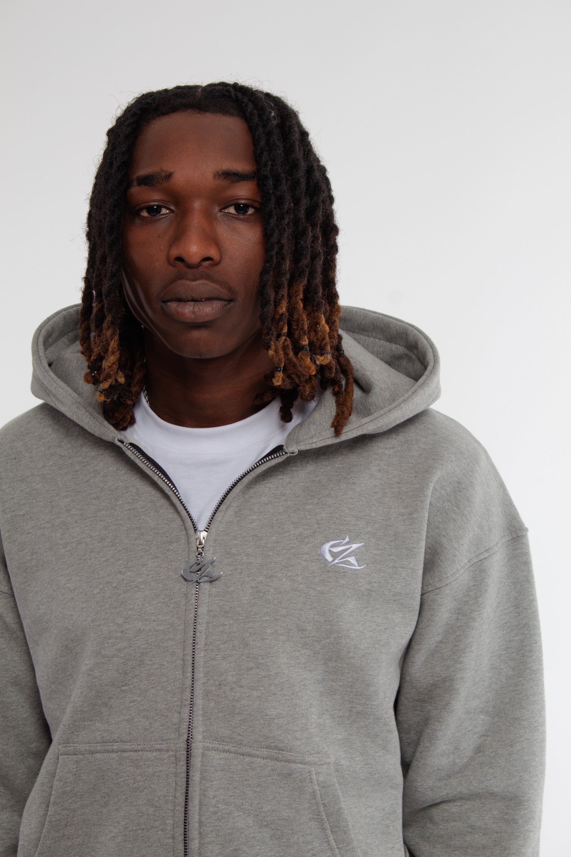 Gros plan sur le hoodie zippé gris Cynique porté, mettant en avant le logo brodé blanc côté cœur. Matière coton 450 GSM, coupe oversize, zip métallique et capuche doublée. Finition premium.