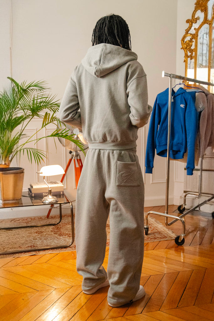 Model wearing Cynique Basic Gray tracksuit from behind — oversized hoodie and joggers in cotton with rear pocket logo, styled in cozy indoor setting