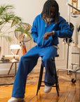 Homme assis portant l’ensemble hoodie zippé et jogging bleu électrique Cynique, coupe oversize unisexe, coton 450 GSM, logo brodé blanc. Look streetwear décontracté dans un intérieur rétro.