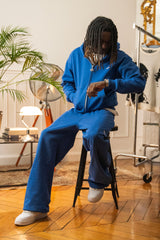 Model seated in full Cynique Basic Blue tracksuit — zip hoodie and baggy joggers styled with white sneakers in a relaxed indoor apartment setting