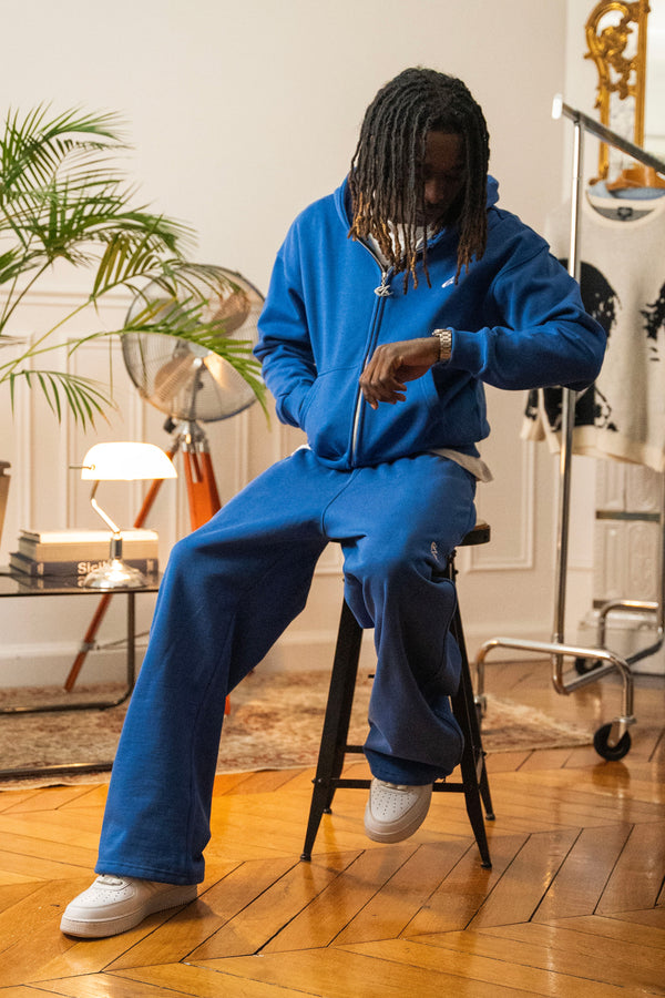 Model seated in full Cynique Basic Blue tracksuit — zip hoodie and baggy joggers styled with white sneakers in a relaxed indoor apartment setting