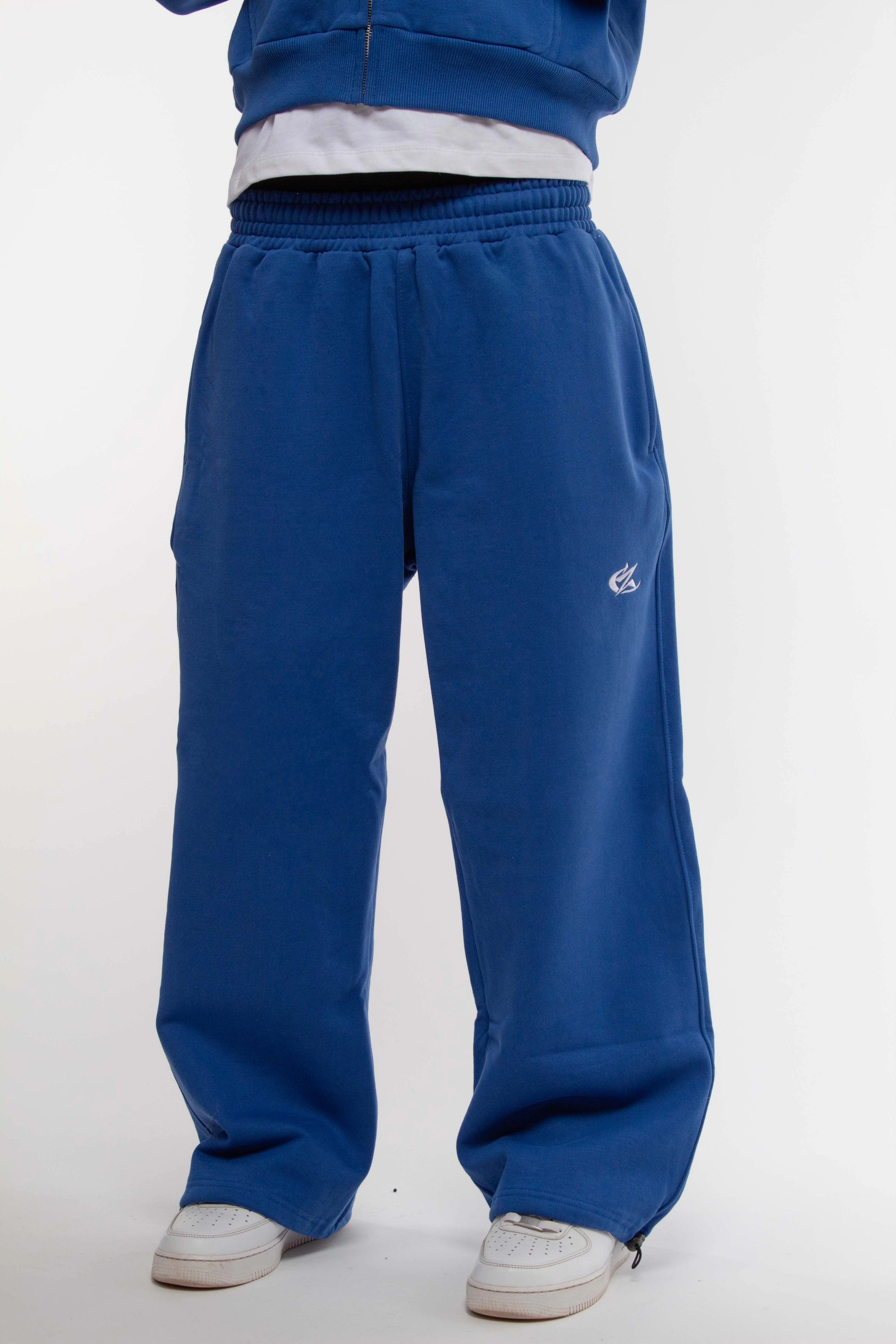 Jogging bleu électrique Cynique en coton épais 450 GSM, coupe oversize avec cordons de serrage aux chevilles. Logo brodé blanc sur la cuisse droite. Porté avec sneakers blanches.
