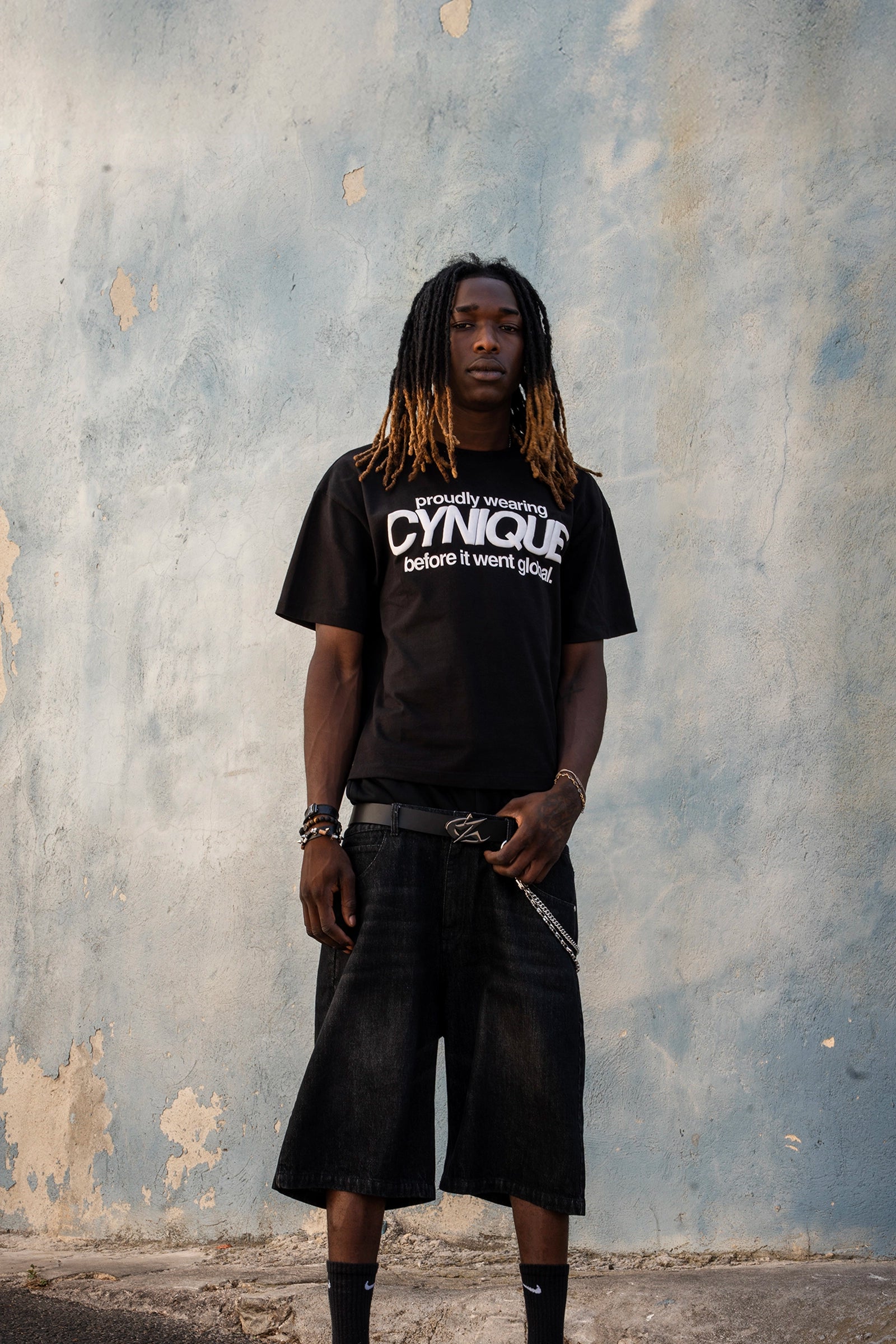 Look streetwear Cynique : T-shirt noir “proudly wearing Cynique before it went global.”, short en denim noir, ceinture logo métal. Photo urbaine sur fond de mur brut texturé.