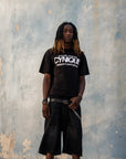 Look streetwear Cynique : T-shirt noir “proudly wearing Cynique before it went global.”, short en denim noir, ceinture logo métal. Photo urbaine sur fond de mur brut texturé.