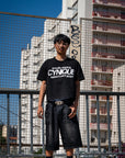 T-shirt streetwear Cynique noir “proudly wearing Cynique before it went global.” et jort denim noir. Look urbain assumé, photo en extérieur devant immeubles et grillage, ambiance raw.