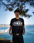 T-shirt noir Cynique “proudly wearing Cynique before it went global.” porté avec jean large, look streetwear minimaliste. Photo en extérieur, ambiance bord de mer.