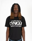 T-shirt noir Cynique “proudly wearing Cynique before it went global.” porté avec jean large bleu et ceinture logo métal. Look streetwear assumé, fond studio blanc.