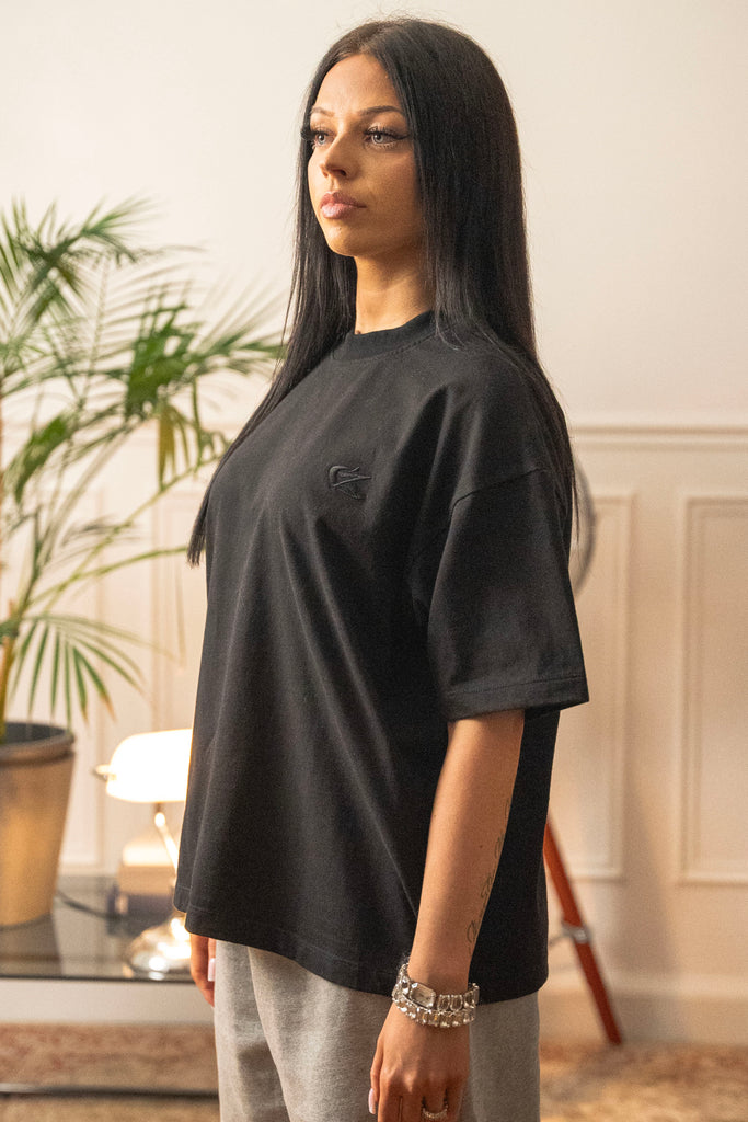 Female model wearing Cynique Vanta Black Tee — oversized unisex T-shirt in 100% cotton with tonal chest logo, styled with gray sweatpants in a relaxed indoor setting