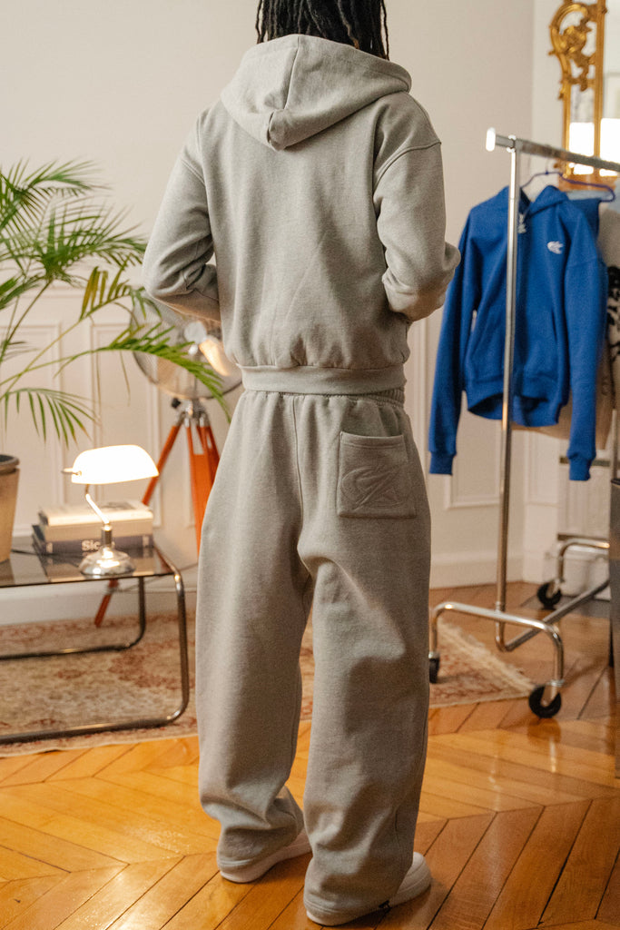Back view of male model wearing Cynique Basic Gray tracksuit — zip hoodie and joggers with tonal embroidered logo on rear pocket, styled in modern apartment setting