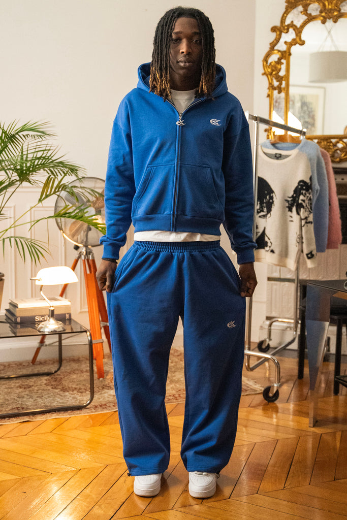 Full-body view of model wearing Cynique Basic Blue tracksuit — oversized hoodie and joggers in 100% cotton, styled with white sneakers in modern interior