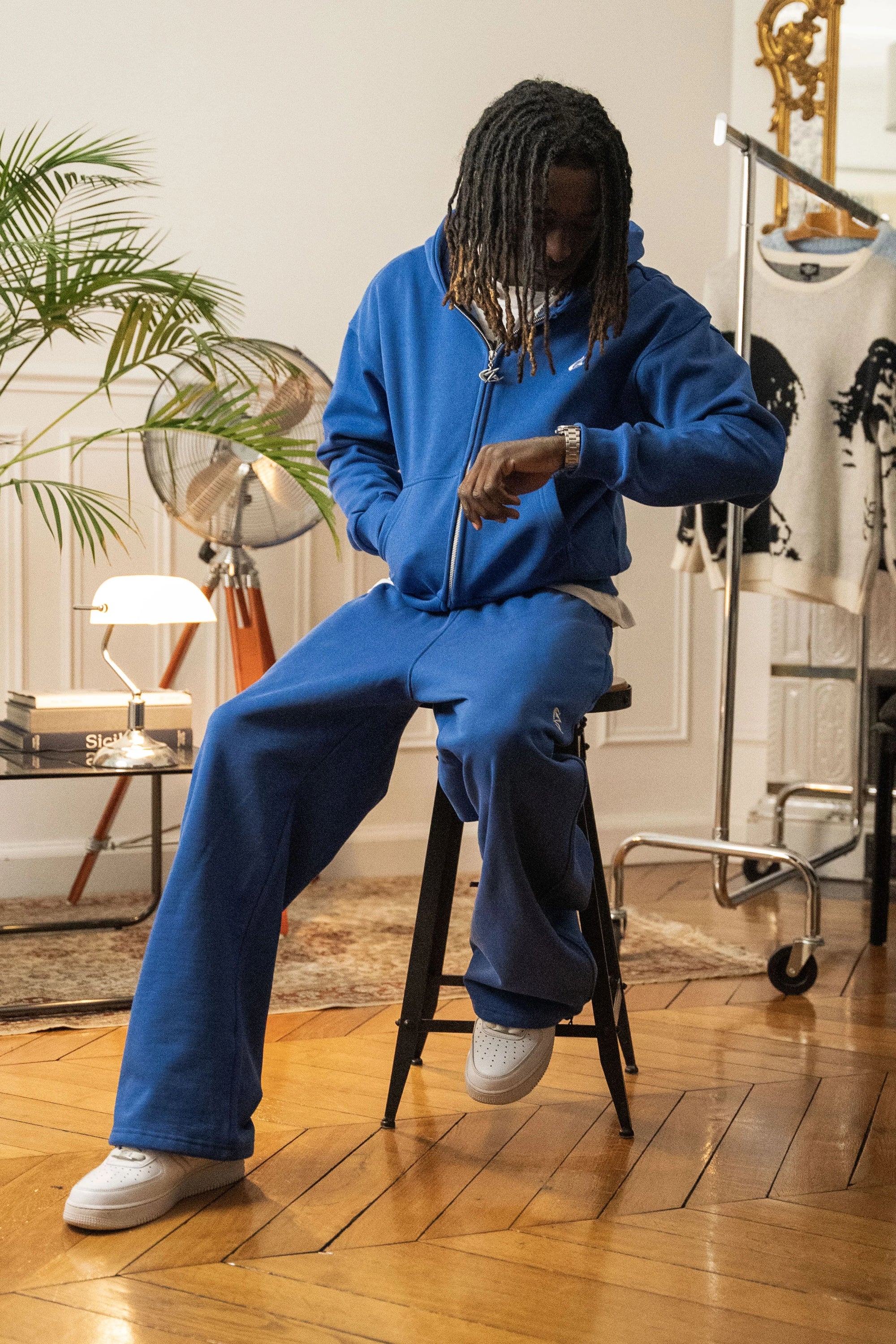 Homme assis sur un tabouret portant un ensemble hoodie zippé et jogging bleu électrique Cynique, coupe oversize, coton 450 GSM, logo brodé blanc, décor rétro intérieur.