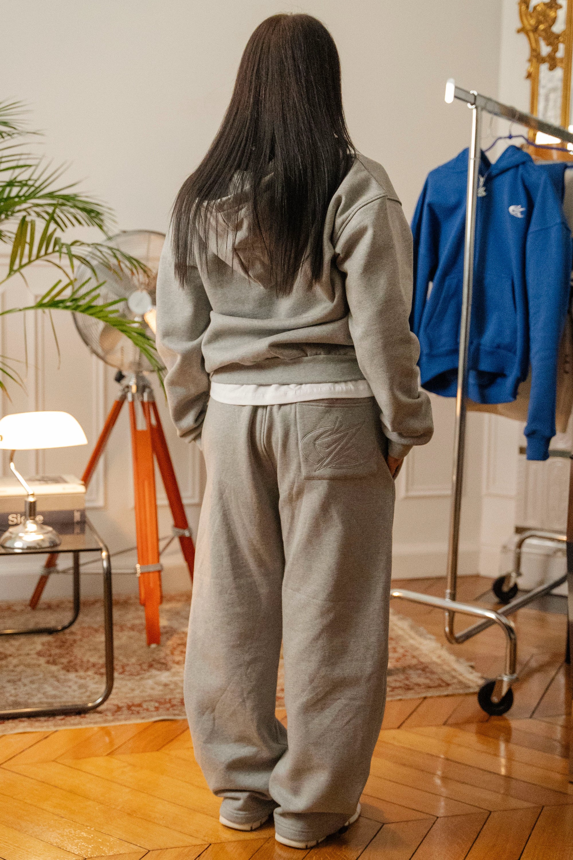 Vue arrière d’un mannequin portant un ensemble hoodie zippé et jogging gris Cynique. Coton 450 GSM, coupe oversize unisexe, poche plaquée arrière, ambiance intérieure stylée.