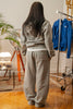 Female model wearing Cynique Basic Gray tracksuit from behind — oversized hoodie and joggers with rear pocket logo, styled in a cozy indoor apartment setting