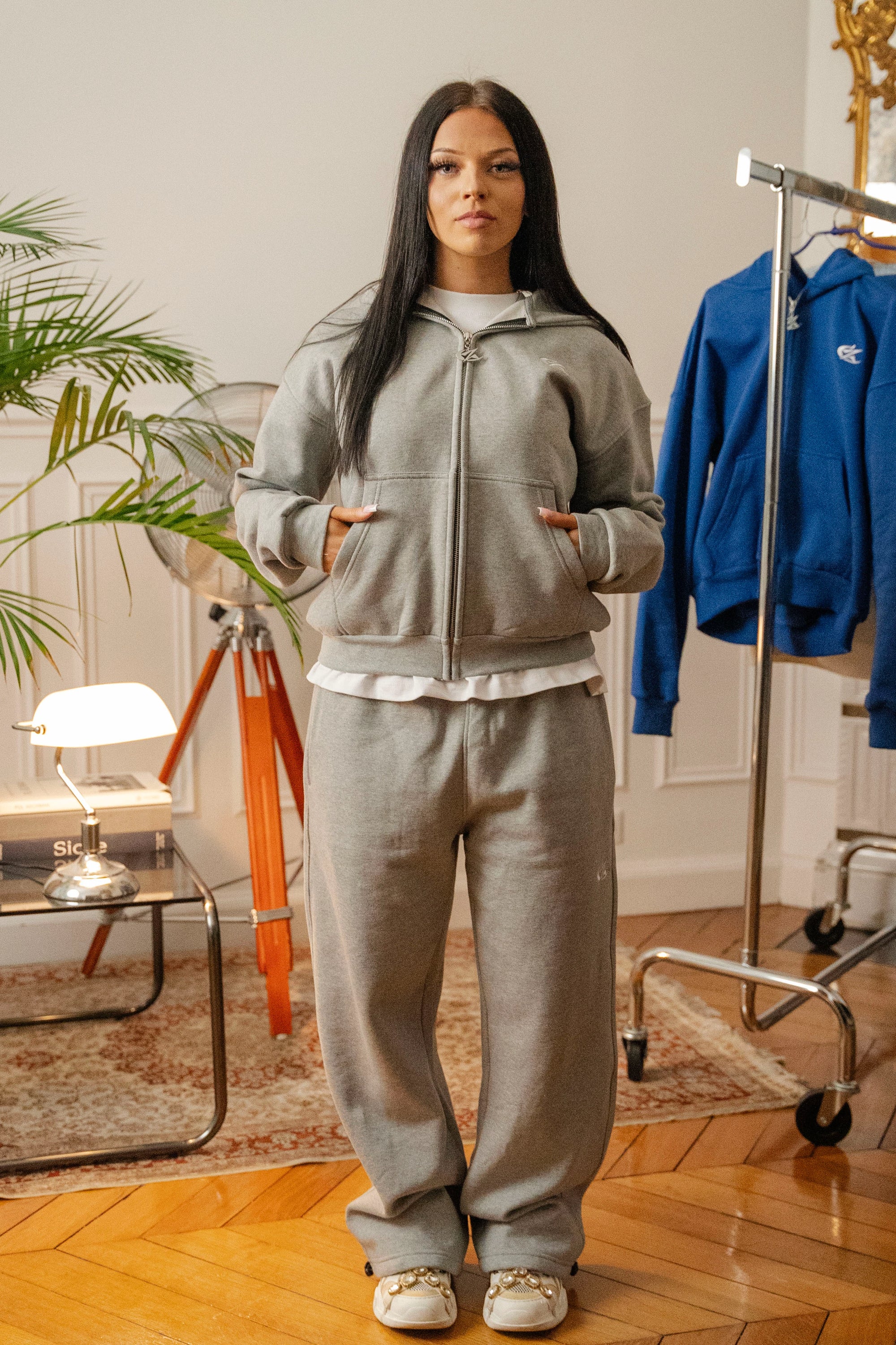 Femme debout portant un ensemble hoodie zippé et jogging gris Cynique. Coupe oversize unisexe, coton 450 GSM, logo brodé blanc, style streetwear épuré dans un intérieur vintage.