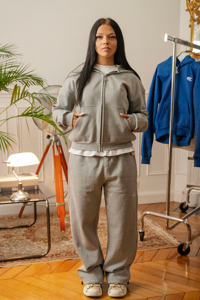 Female model wearing Cynique Basic Gray tracksuit — boxy zip-up hoodie and oversized joggers in soft cotton, styled with statement sneakers in a modern interior