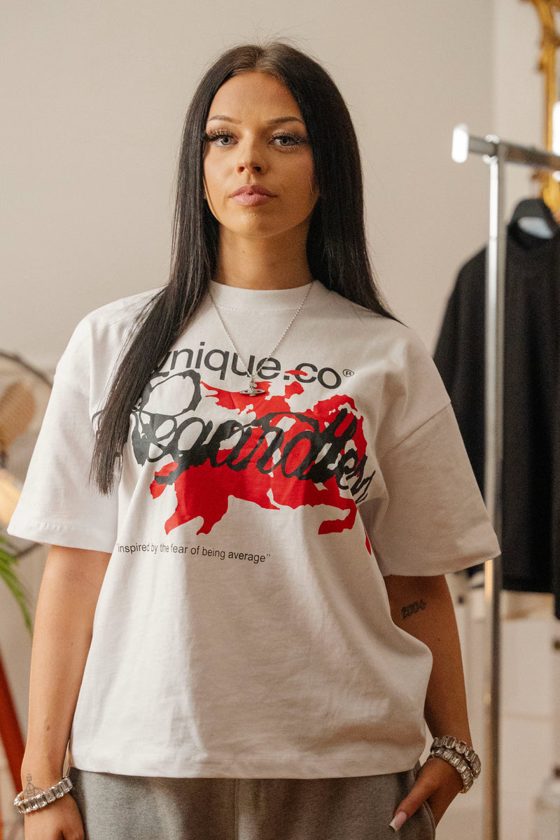 T-shirt blanc Cynique porté par une femme. Design streetwear avec visuel “Regardless” noir et rouge imprimé sur la poitrine, slogan “inspired by the fear of being average”. Coupe oversize unisexe, coton 210 GSM. Look urbain et affirmé.