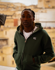 Hoodie Cynique vert porté, ambiance ocre de la médina. Coupe large, broderie blanche discrète, pièce essentielle pour un style streetwear premium et décontracté.